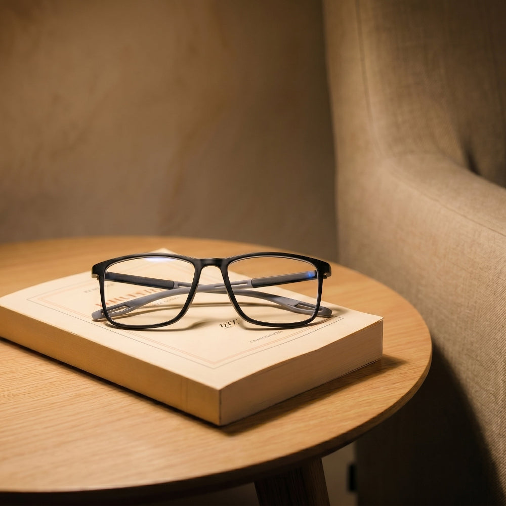 Lunettes de lecture à monture grise posées pliées sur un livre ouvert retourné, table de chevet en bois clair, lumière chaude de lampe de chevet, verres transparents laissant voir les pages ivoire Image